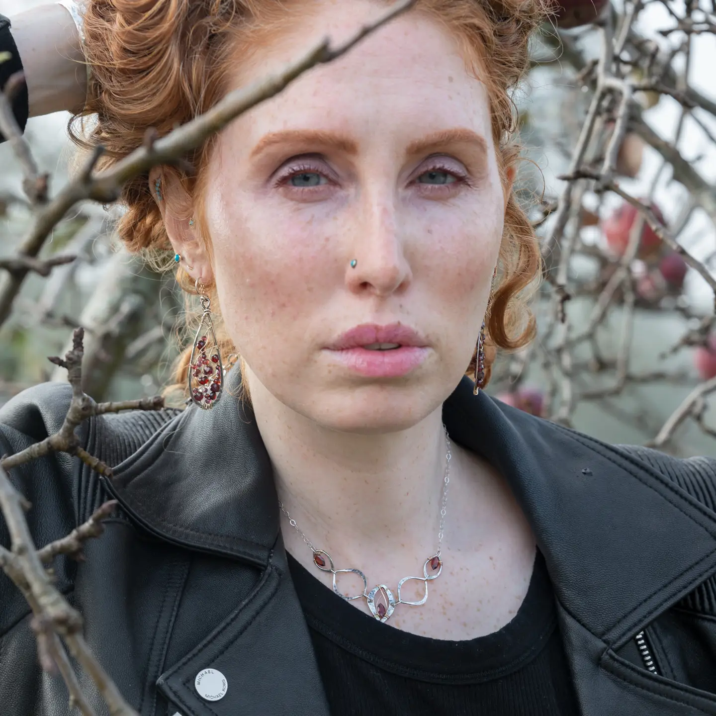 Woman with red hair and freckles standing among tree branches wearing garnet earrings.
