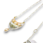 A delicate necklace featuring a crescent-shaped pendant with white freshwater pearls, a light green stone, and a pearlescent gold-toned accent, suspended on a silver chain against a white background.