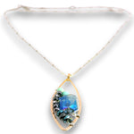 A gold teardrop pendant necklace featuring a central blue iridescent stone and black beads, suspended from a white cord on a white background.