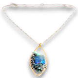 A gold teardrop pendant necklace featuring a central blue iridescent stone and black beads, suspended from a white cord on a white background.