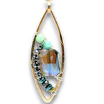 A gold-toned teardrop pendant featuring a large faceted iridescent blue gemstone, accented with light green beads, black faceted beads, and smaller blue-green beads on a white background.