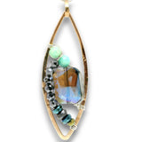 A gold-toned teardrop pendant featuring a large faceted iridescent blue gemstone, accented with light green beads, black faceted beads, and smaller blue-green beads on a white background.