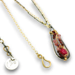A close-up of a necklace with a teardrop-shaped pendant featuring a silver-toned textured frame, gold-toned wire wrapping, and a cluster of pink and burgundy gemstones. A gold-toned chain and a small round silver-toned charm engraved with a symbol are also visible against a white background.