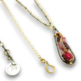 A close-up of a necklace with a teardrop-shaped pendant featuring a silver-toned textured frame, gold-toned wire wrapping, and a cluster of pink and burgundy gemstones. A gold-toned chain and a small round silver-toned charm engraved with a symbol are also visible against a white background.