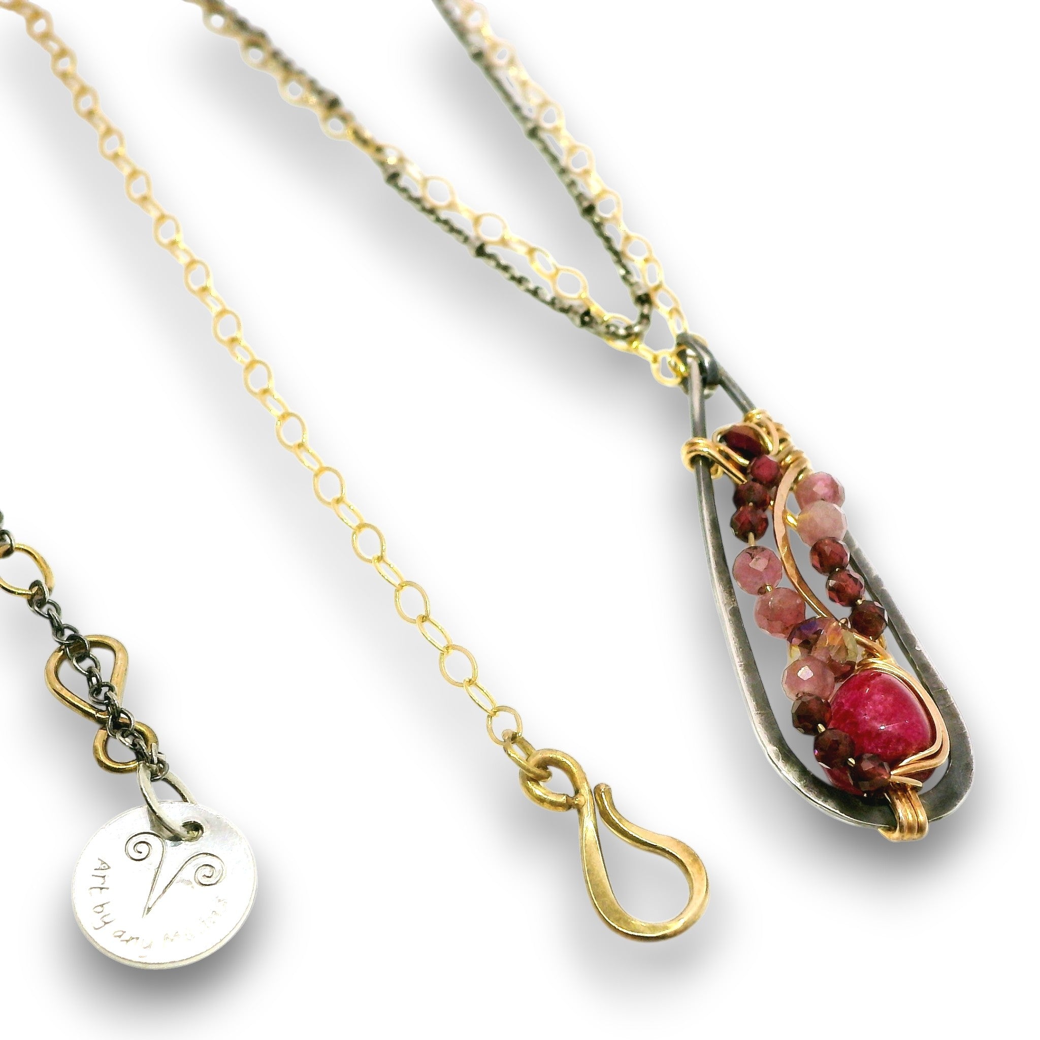 A close-up of a necklace with a teardrop-shaped pendant featuring a silver-toned textured frame, gold-toned wire wrapping, and a cluster of pink and burgundy gemstones. A gold-toned chain and a small round silver-toned charm engraved with a symbol are also visible against a white background.