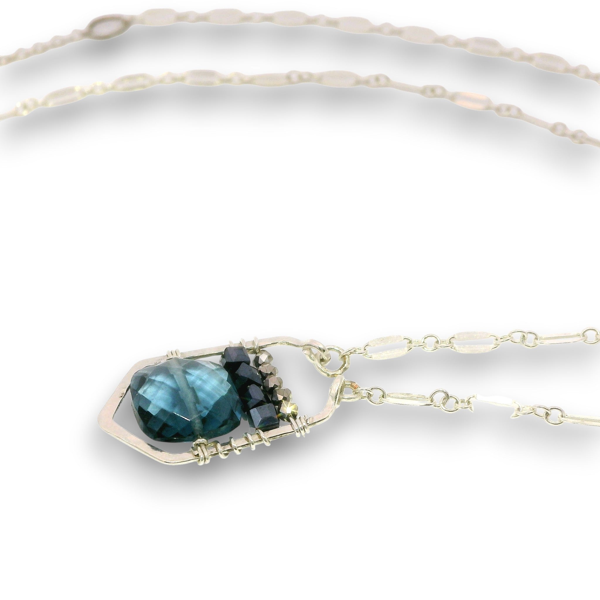 A silver necklace featuring a unique pendant with a faceted dark blue-green gemstone wrapped in silver wire, accented with small dark blue and clear crystals, shown on a white background.