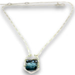 A silver-toned necklace featuring a textured chain and a central pendant with a faceted dark teal-blue gemstone held in a silver setting with small dark accent stones, shown on a white background.