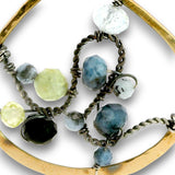 A close-up of a gold-tone and dark bronze wire-wrapped necklace featuring faceted gemstones in shades of blue, pale yellow-green, black, and white, set against a plain white background.