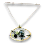 A gold-toned pendant necklace featuring a large open oval frame with a white cord and adorned with faceted beads in black, light green, and blue, set against a plain white background.