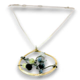 A gold-toned pendant necklace featuring a large open oval frame with a white cord and adorned with faceted beads in black, light green, and blue, set against a plain white background.