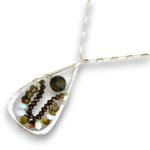 A teardrop-shaped silver pendant necklace featuring a large round tiger's eye stone and two strands of faceted beads in brown, olive green, and white, suspended from a silver chain on a white background.
