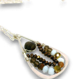 A close-up of a teardrop-shaped pendant necklace featuring a silver-toned frame, a dark brown tiger's eye bead, and multiple strands of faceted beads in brown, gold, and pale blue, set against a plain white background.