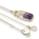 A gold chain necklace with a rectangular pendant featuring a polished purple amethyst crystal, wrapped with delicate gold wire, shown on a white background with the adjustable chain and clasp visible.