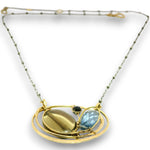 A gold-toned celestial pendant necklace featuring a large oval gold cabochon, a faceted light blue teardrop gemstone, a small dark round gemstone, and a delicate beaded chain.