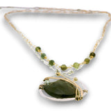 A beaded necklace featuring a large oval green stone pendant wrapped in silver and gold wire, accented with small green beads and clear crystal beads on a white background.