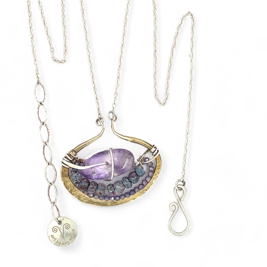 A sterling silver adjustable chain necklace with a hammered handmade brass pendant featuring a glowing amethyst smooth pebble, surrounded by faceted blue zoisite and micro faceted lavender cubic zirconia gemstones.