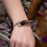 A handcrafted sterling silver bracelet featuring a faceted drop of dark amethyst and other amethyst gemstones in hammered brass, accompanied by micro-faceted cubic zirconia