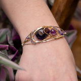 A handcrafted sterling silver bracelet featuring a faceted drop of dark amethyst and other amethyst gemstones in hammered brass, accompanied by micro-faceted cubic zirconia