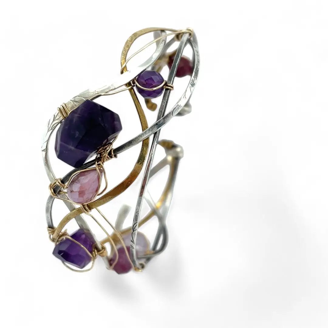 A collection of faceted amethyst nuggets, pink sapphire, and ruby gemstones are wrapped in gold filled wire onto a bracelet formed of leaf stamped oxidized sterling, bright sterling, and bronze hammered waves