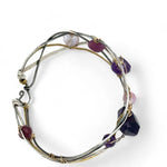 A collection of faceted amethyst nuggets, pink sapphire, and ruby gemstones are wrapped in gold filled wire onto a bracelet formed of leaf stamped oxidized sterling, bright sterling, and bronze hammered waves