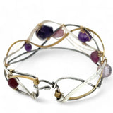 A collection of faceted amethyst nuggets, pink sapphire, and ruby gemstones are wrapped in gold filled wire onto a bracelet formed of leaf stamped oxidized sterling, bright sterling, and bronze hammered waves