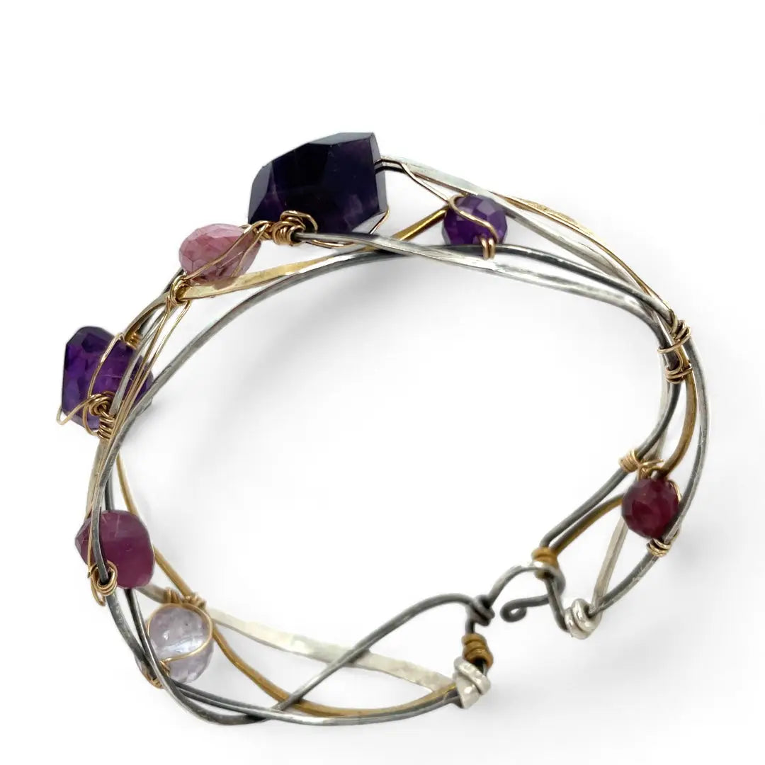 A collection of faceted amethyst nuggets, pink sapphire, and ruby gemstones are wrapped in gold filled wire onto a bracelet formed of leaf stamped oxidized sterling, bright sterling, and bronze hammered waves