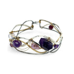 A collection of faceted amethyst nuggets, pink sapphire, and ruby gemstones are wrapped in gold filled wire onto a bracelet formed of leaf stamped oxidized sterling, bright sterling, and bronze hammered waves