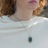 A sterling silver necklace featuring a large, oval-shaped teal apatite gemstone encircled by smaller faceted chrysocolla gemstones, set in gold-filled and bronze accents, displayed on a silver chain.