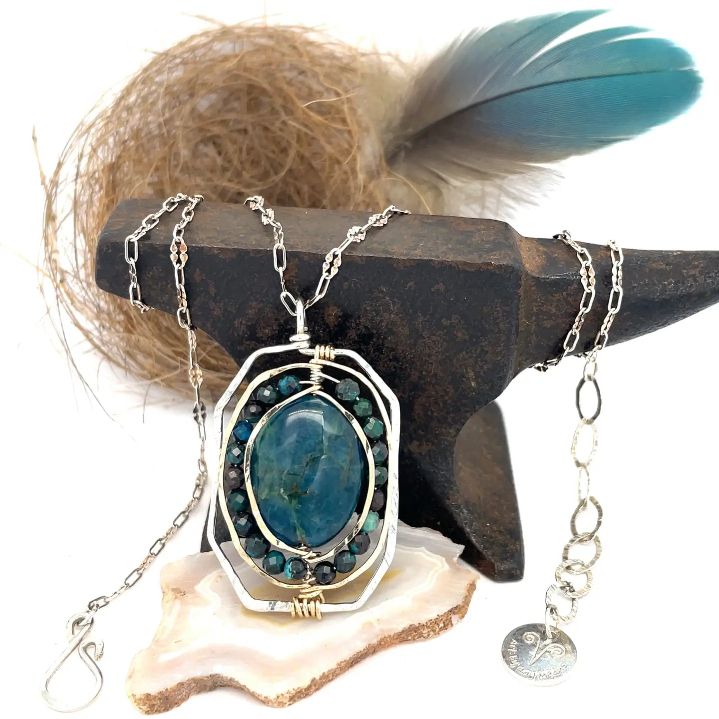 A sterling silver necklace featuring a large, oval-shaped teal apatite gemstone encircled by smaller faceted chrysocolla gemstones, set in gold-filled and bronze accents, displayed on a silver chain.