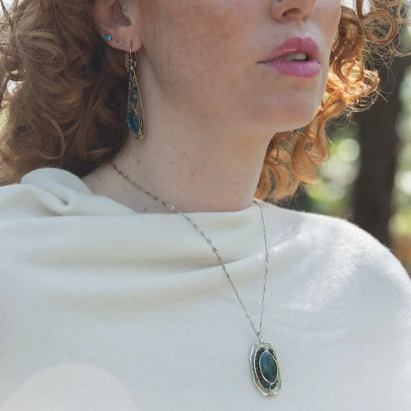 A sterling silver necklace featuring a large, oval-shaped teal apatite gemstone encircled by smaller faceted chrysocolla gemstones, set in gold-filled and bronze accents, displayed on a silver chain.