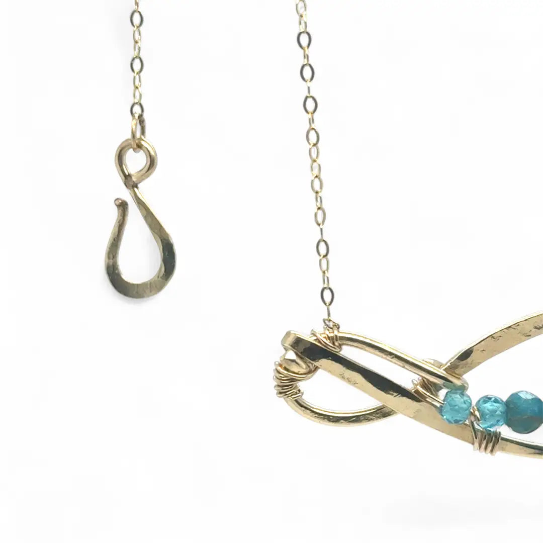 A brass infinity-shaped pendant necklace with blue faceted apatite and opal gemstones on the inside, attached to a gold-filled chain with an adjustable clasp.