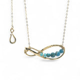 A brass infinity-shaped pendant necklace with blue faceted apatite and opal gemstones on the inside, attached to a gold-filled chain with an adjustable clasp.