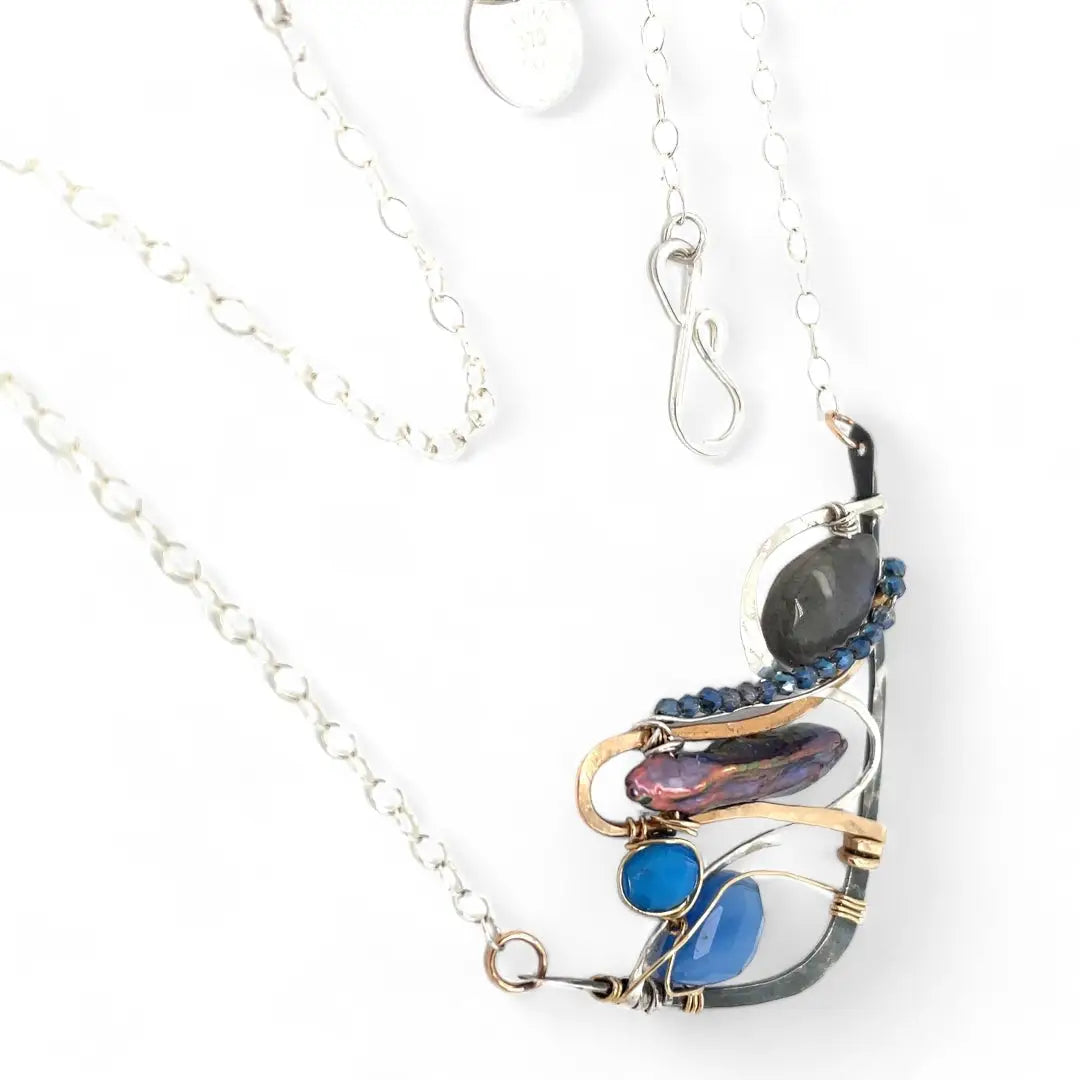 Darkened sterling silver boomerang shaped pendant necklace with waves of hammered bronze, sterling silver, and gold filled wire holding smooth labradorite, blue jade, blue crystal, and a purple fresh water stick pearl in place