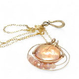 A gold-filled chain necklace with a pearl pink pearl coin inside a bronze ring wrapped in gold-filled rings with a layer of peach cat's eye and pink opal gemstones
