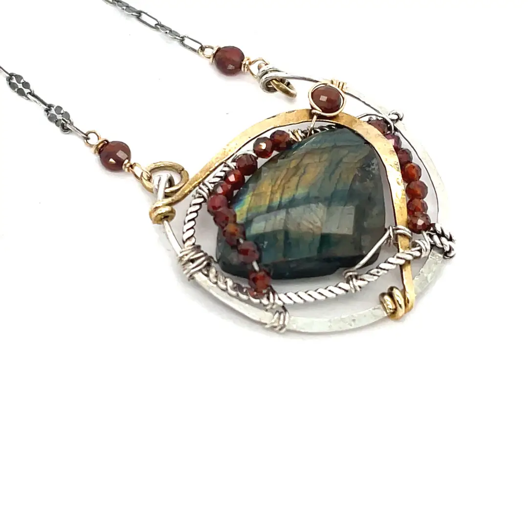 A necklace with teardrop-shaped labradorite gemstone adorned with faceted hessonite gemstones and set between hammered arcs of brass and oxidized sterling twist inside a sterling silver semi circle pendant. 