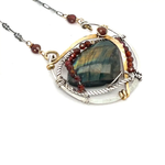 A necklace with teardrop-shaped labradorite gemstone adorned with faceted hessonite gemstones and set between hammered arcs of brass and oxidized sterling twist inside a sterling silver semi circle pendant. 