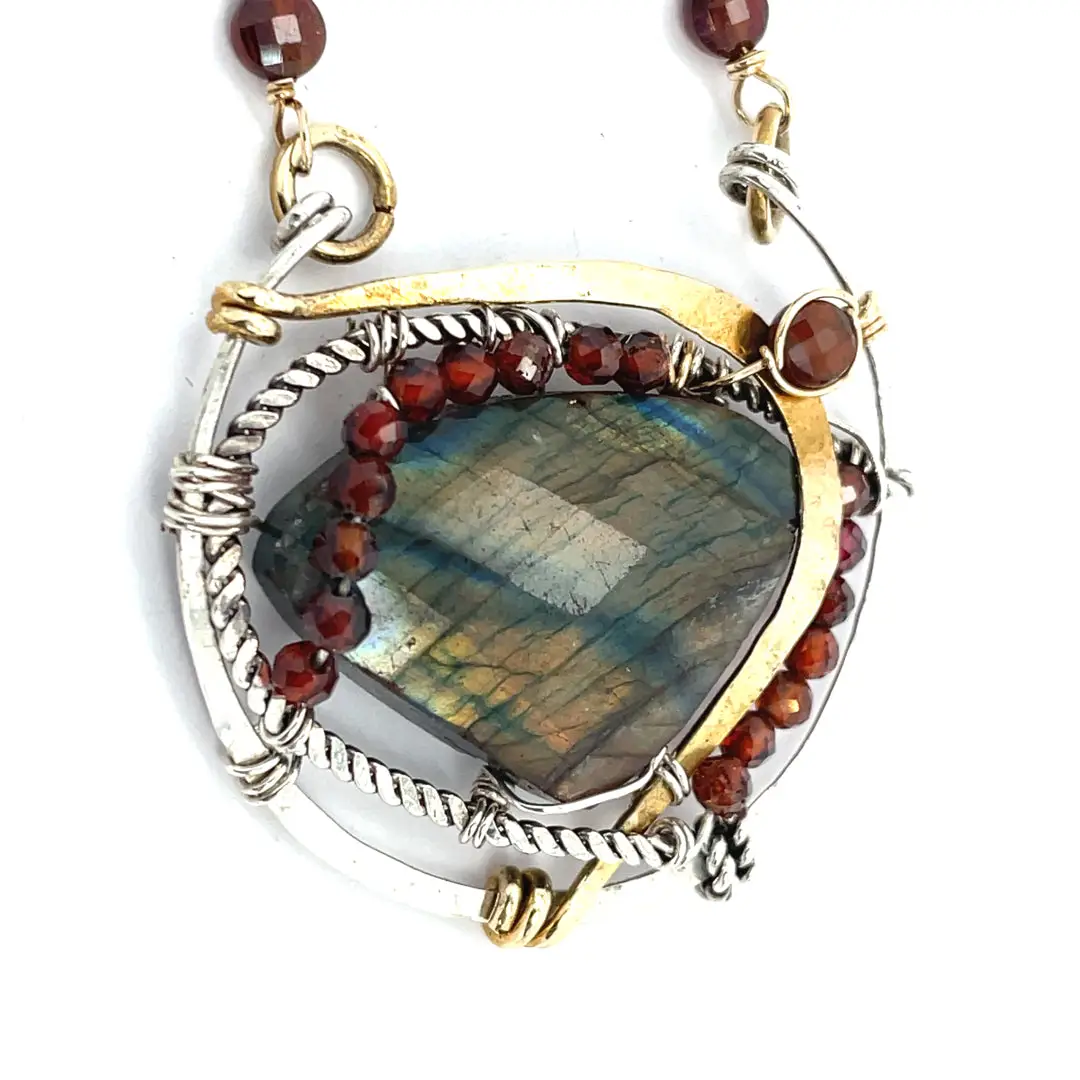 A necklace with teardrop-shaped labradorite gemstone adorned with faceted hessonite gemstones and set between hammered arcs of brass and oxidized sterling twist inside a sterling silver semi circle pendant. 