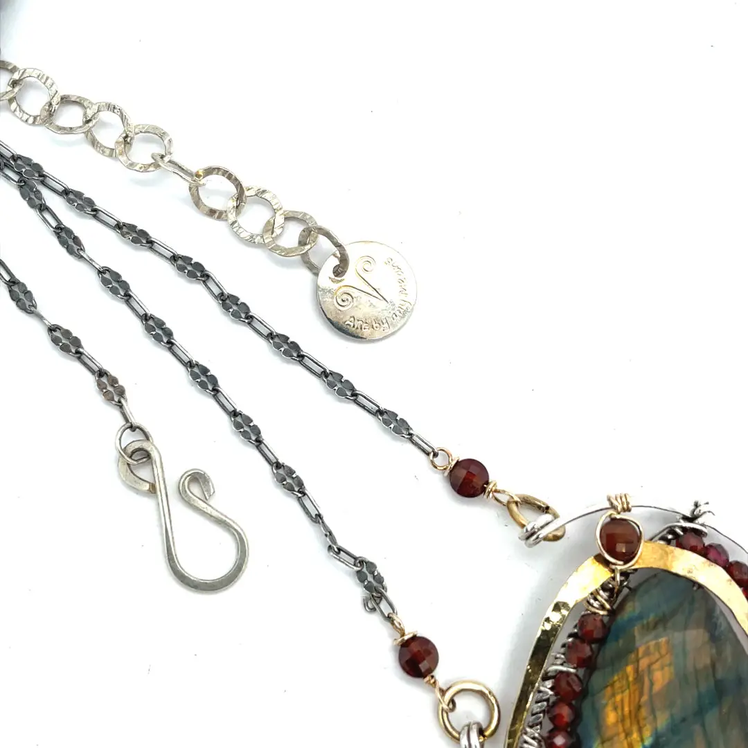 A necklace with teardrop-shaped labradorite gemstone adorned with faceted hessonite gemstones and set between hammered arcs of brass and oxidized sterling twist inside a sterling silver semi circle pendant. 