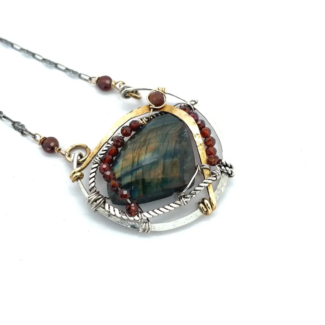 A necklace with teardrop-shaped labradorite gemstone adorned with faceted hessonite gemstones and set between hammered arcs of brass and oxidized sterling twist inside a sterling silver semi circle pendant. 