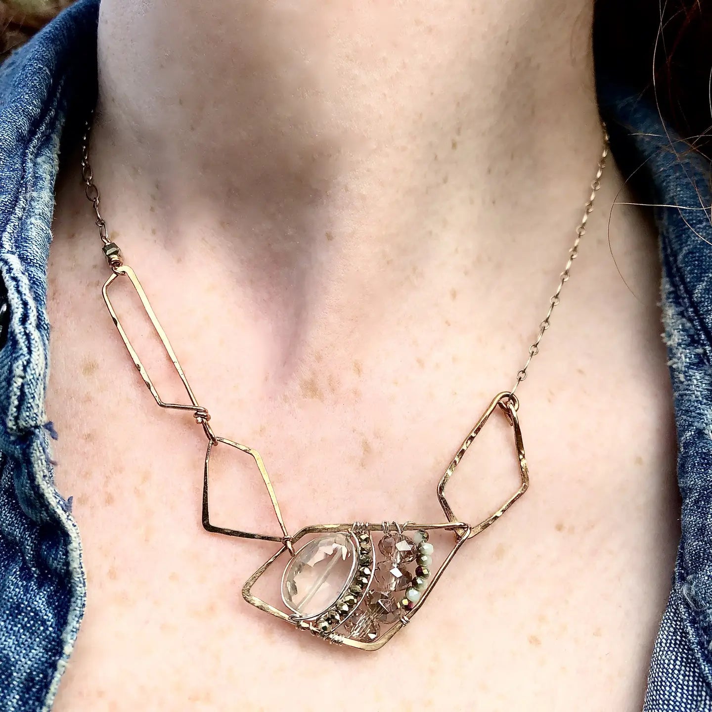 A faceted clear quartz oval gemstone is layered with pyrite and crystals on sterling silver wire inside a bronze parallelogram, then attached to three varying sized parallelograms