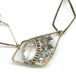 A faceted clear quartz oval gemstone is layered with pyrite and crystals on sterling silver wire inside a bronze parallelogram, then attached to three varying sized parallelograms