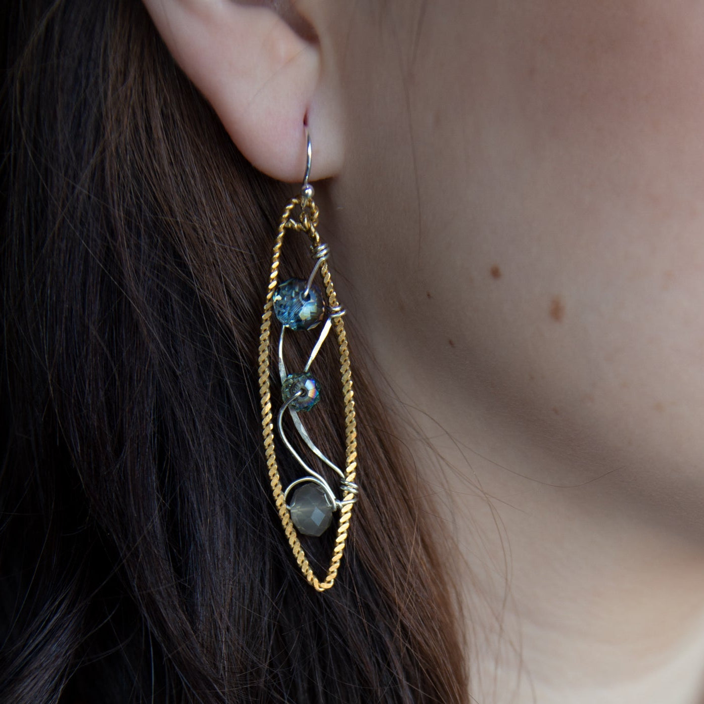 A pair of handmade brass twisted marquise earrings with hammered sterling silver and sparkling crystals in blue, green, and grey colors.
