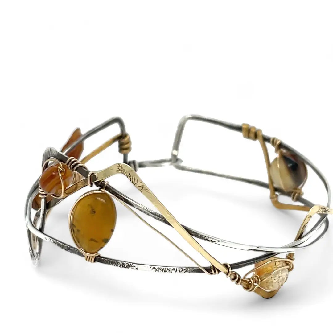 A handcrafted cuff bracelet featuring a mix of yellow opal, faceted citrine, banded agate, and hessonite raw crystal, wrapped in gold fill and set in among brass and oxidized sterling silver geometric cuff bracelet that's been stamped with trees