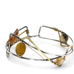 A handcrafted cuff bracelet featuring a mix of yellow opal, faceted citrine, banded agate, and hessonite raw crystal, wrapped in gold fill and set in among brass and oxidized sterling silver geometric cuff bracelet that's been stamped with trees