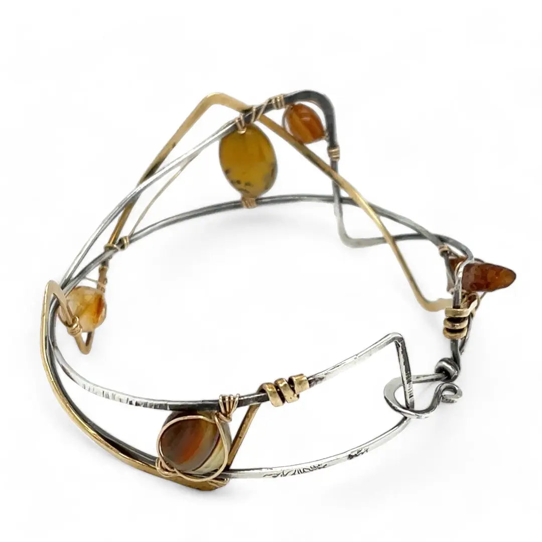 A handcrafted cuff bracelet featuring a mix of yellow opal, faceted citrine, banded agate, and hessonite raw crystal, wrapped in gold fill and set in among brass and oxidized sterling silver geometric cuff bracelet that's been stamped with trees