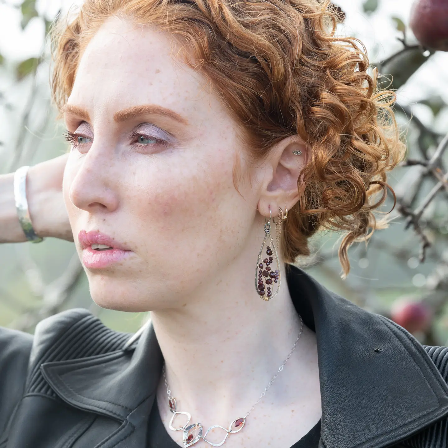 Woman with curly red hair wearing earrings and a necklace, outdoors.