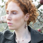 Woman with curly red hair wearing earrings and a necklace, outdoors.