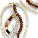 A close-up of a beaded jewelry piece featuring faceted beads in reddish-brown, white, peach, yellow, and olive green, suspended on a textured gold and silver-tone metal frame.