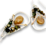 A pair of handmade dangle earrings featuring a large smooth, polished tan stone in a silver-toned frame, accented with dark green and bronze beads, on a white background.
