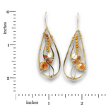 A pair of gold teardrop dangle earrings with amber and glass bead details, shown next to a black measurement ruler on a white background.